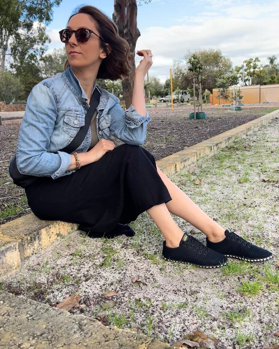 Woman sitting outdoors wearing a denim jacket and black shoes.
